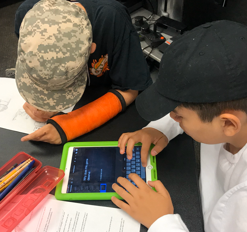 Two students at a table working on a project — one writing and one using a tablet